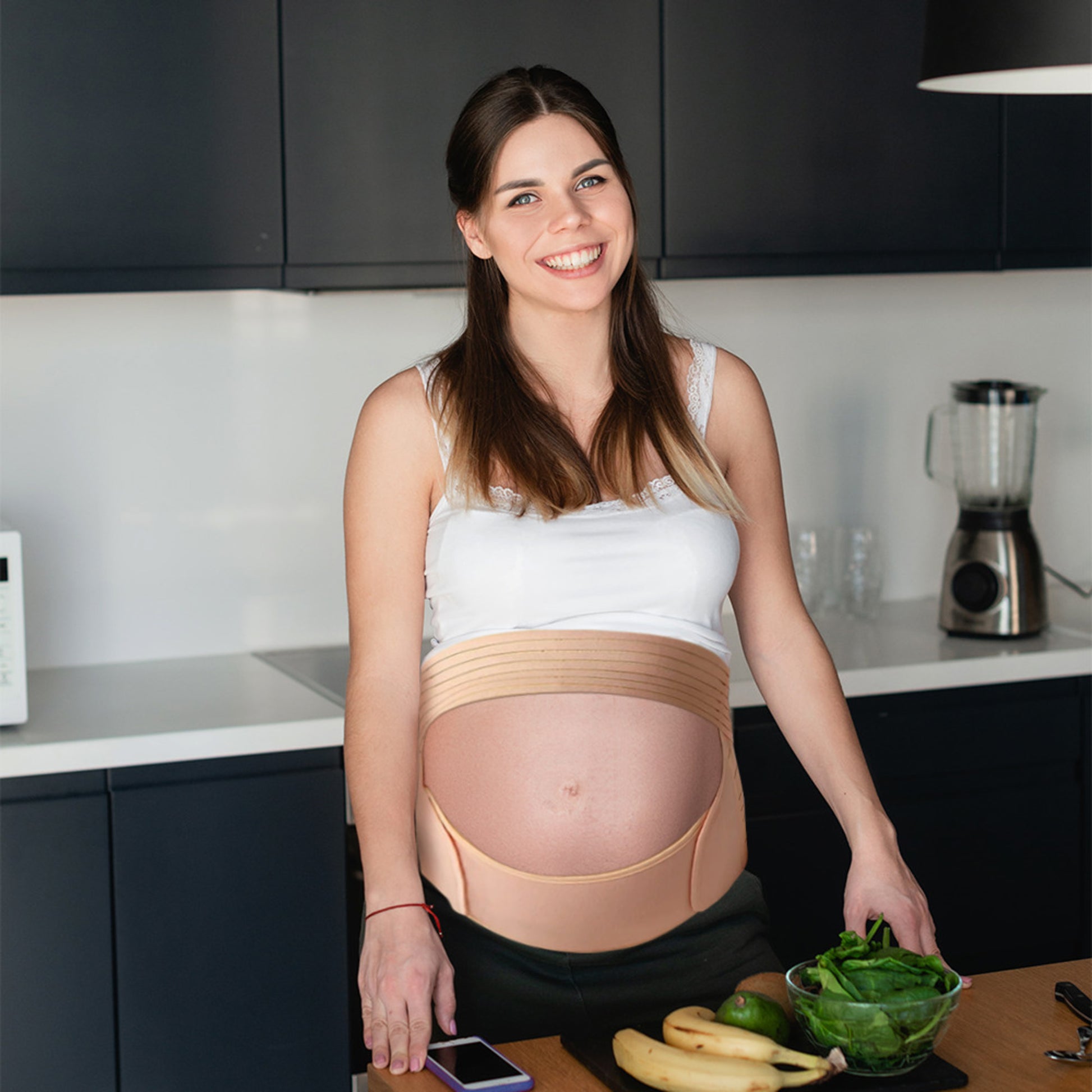 Gravid kvinne som står på kjøkkenet med Luumii ComfortBump støttebelte i beige for avlastning av mage og rygg

